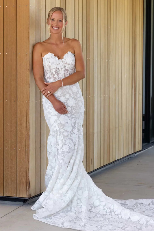 A woman wearing a strapless white lace wedding gown with a sweetheart neckline and a long train, standing against a light wood-paneled wall.