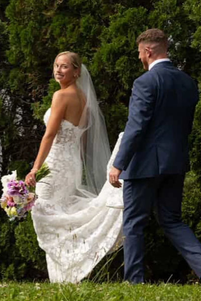 A woman wearing a strapless white lace wedding gown with a sweetheart neckline and a long train, standing against a light wood-paneled wall.