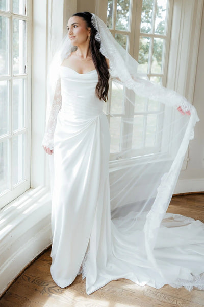 A woman wearing a white off-the-shoulder wedding gown with long lace sleeves and a long sheer veil, standing in a sunlit room with large windows and wood floors.