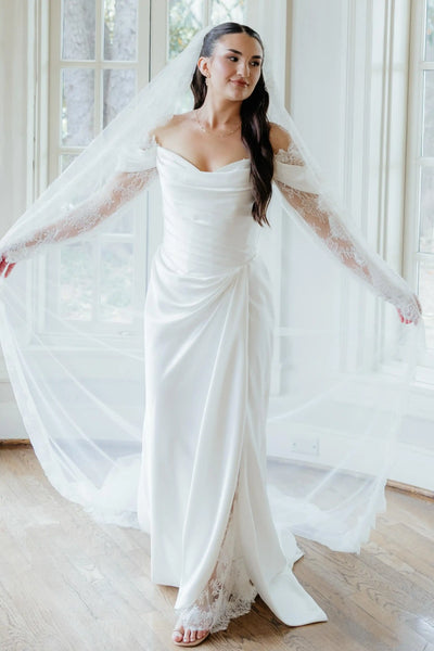 A woman wearing a white off-the-shoulder wedding gown with long lace sleeves and a long sheer veil, standing in a sunlit room with large windows and wood floors.