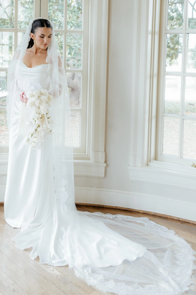 A woman wearing a white off-the-shoulder wedding gown with long lace sleeves and a long sheer veil, standing in a sunlit room with large windows and wood floors.