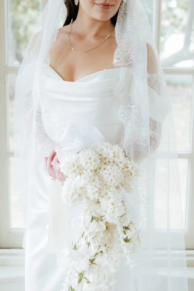 A woman wearing a white off-the-shoulder wedding gown with long lace sleeves and a long sheer veil, standing in a sunlit room with large windows and wood floors.