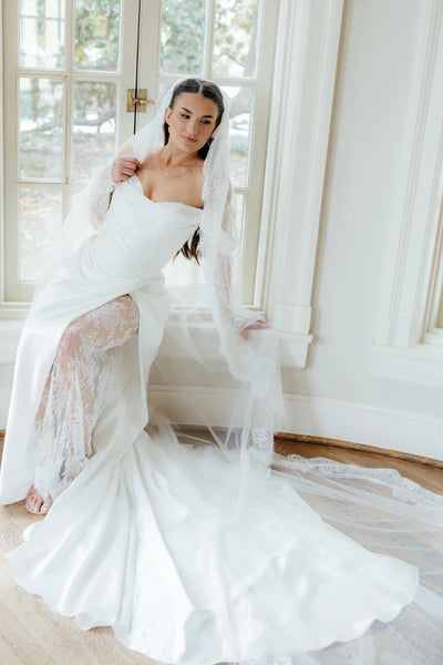 A woman wearing a white off-the-shoulder wedding gown with long lace sleeves and a long sheer veil, standing in a sunlit room with large windows and wood floors.