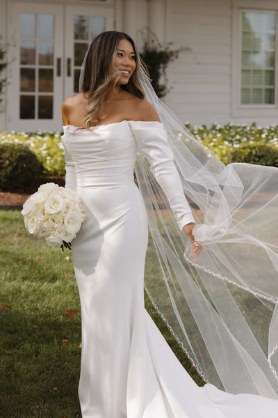 A woman wearing an off-the-shoulder white satin wedding gown with long sleeves and a long sheer tulle veil, standing outdoors in front of a house and holding a bouquet of white roses.