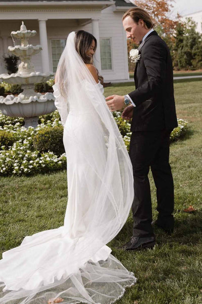 A woman wearing an off-the-shoulder white satin wedding gown with long sleeves and a long sheer tulle veil, standing outdoors in front of a house and holding a bouquet of white roses.