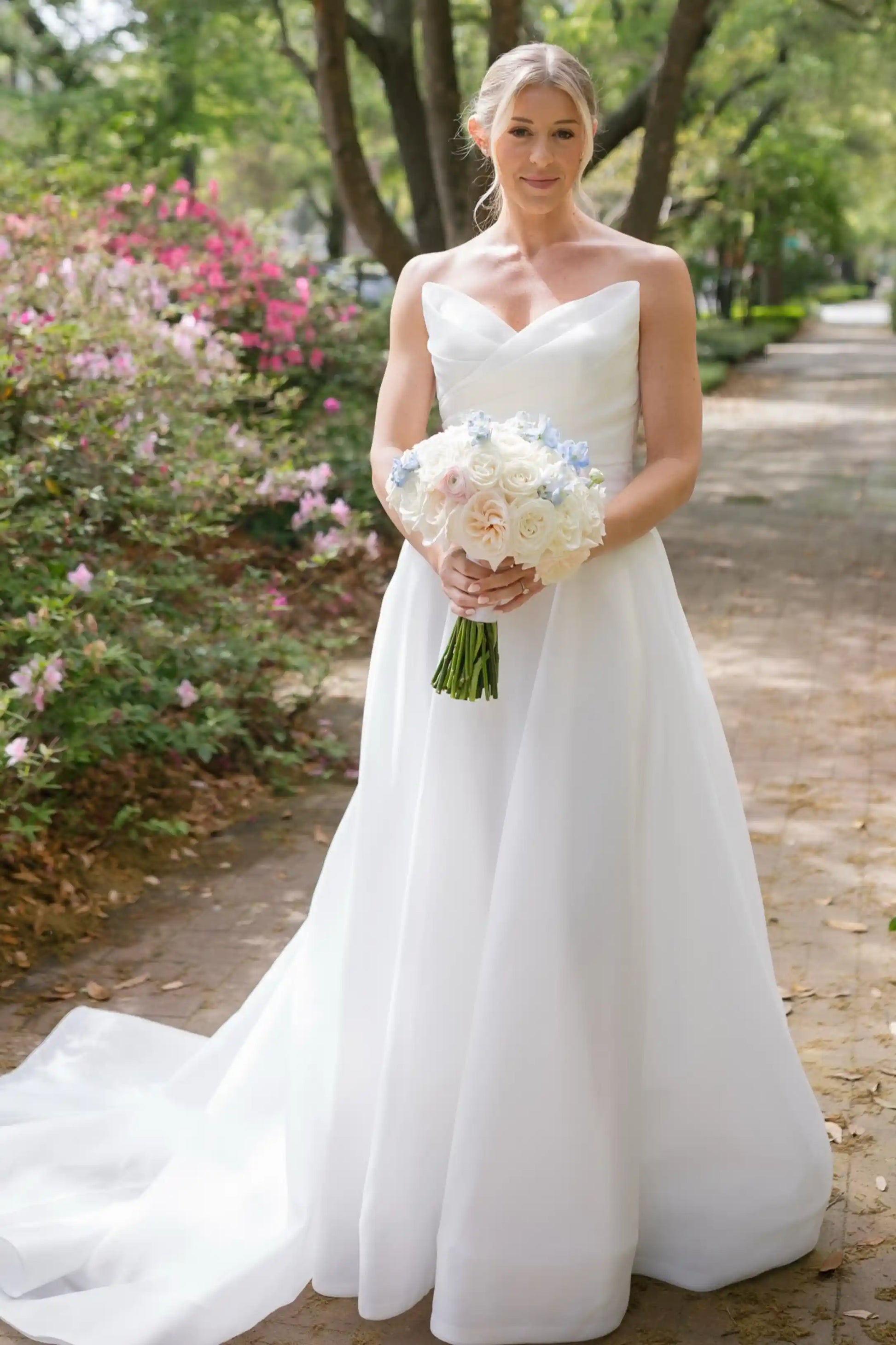 A woman wearing a strapless white wedding gown stands on a tree-lined path holding a bouquet of white, cream, and light blue flowers.