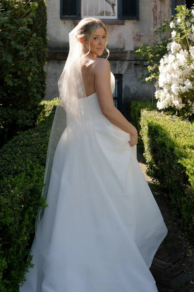 A woman wearing a strapless white wedding gown stands on a tree-lined path holding a bouquet of white, cream, and light blue flowers.