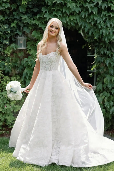 A woman wearing a white lace wedding gown with a long sheer veil and holding a bouquet of white flowers, standing outdoors in front of a leafy green wall.