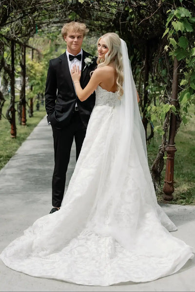 A woman wearing a white lace wedding gown with a long sheer veil and holding a bouquet of white flowers, standing outdoors in front of a leafy green wall.