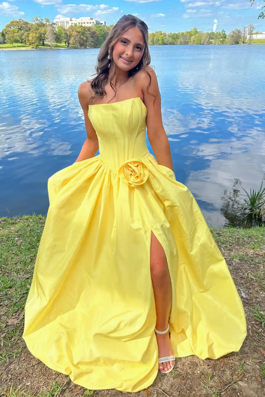 A young woman wearing  yellow strapless ball gown with a large fabric rose detail at the waist and a high leg slit, standing outdoors on a grassy bank with a lake and trees in the background.
