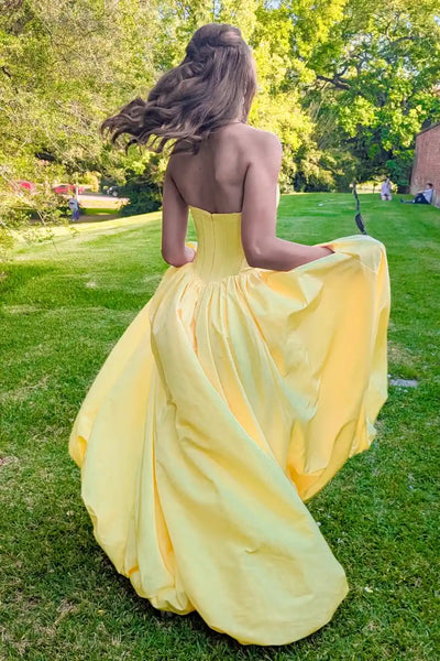 A young woman wearing  yellow strapless ball gown with a large fabric rose detail at the waist and a high leg slit, standing outdoors on a grassy bank with a lake and trees in the background.
