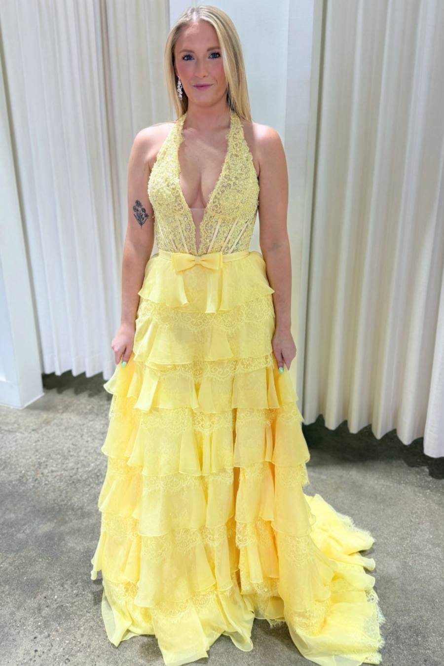 A woman wearing a bright yellow formal gown with a lace halter neckline, plunging V-neckline, and a tiered ruffled skirt, standing in front of a white curtain.
