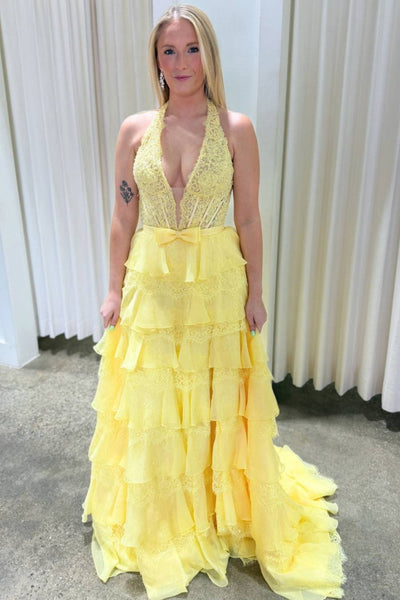A woman wearing a bright yellow formal gown with a lace halter neckline, plunging V-neckline, and a tiered ruffled skirt, standing in front of a white curtain.
