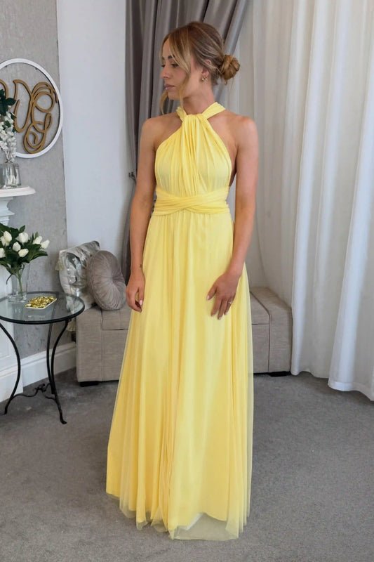 A woman wearing a yellow halter-neck maxi dress with a pleated bodice and flowing tulle skirt, standing in a living room setting.
