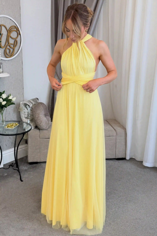 A woman wearing a yellow halter-neck maxi dress with a gathered waist, standing in a living room setting with curtains, a sofa, and a side table.
