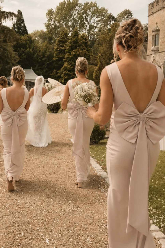 Bridesmaids in matching satin dresses with large bow back details walk down a gravel path toward a bride in a white wedding gown in an outdoor garden setting.