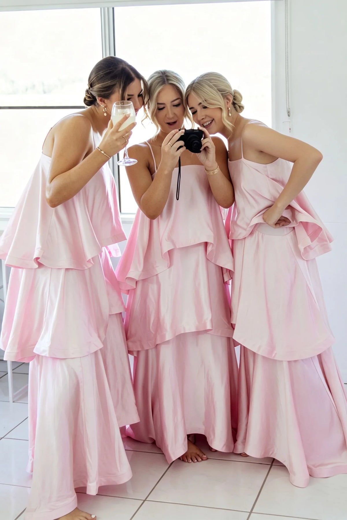 Three women wearing matching light pink tiered satin bridesmaid dresses pose together in a bright room, with one holding a camera and another holding a glass.