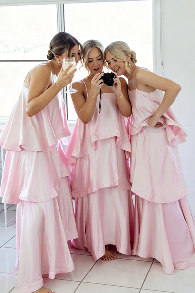 Three women wearing matching light pink tiered satin bridesmaid dresses pose together in a bright room, with one holding a camera and another holding a glass.