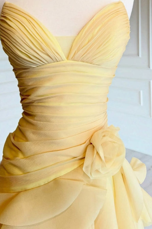A yellow strapless gown with a sweetheart neckline, pleated bodice, ruched waist, and a large fabric flower detail on the skirt, displayed on a mannequin torso.