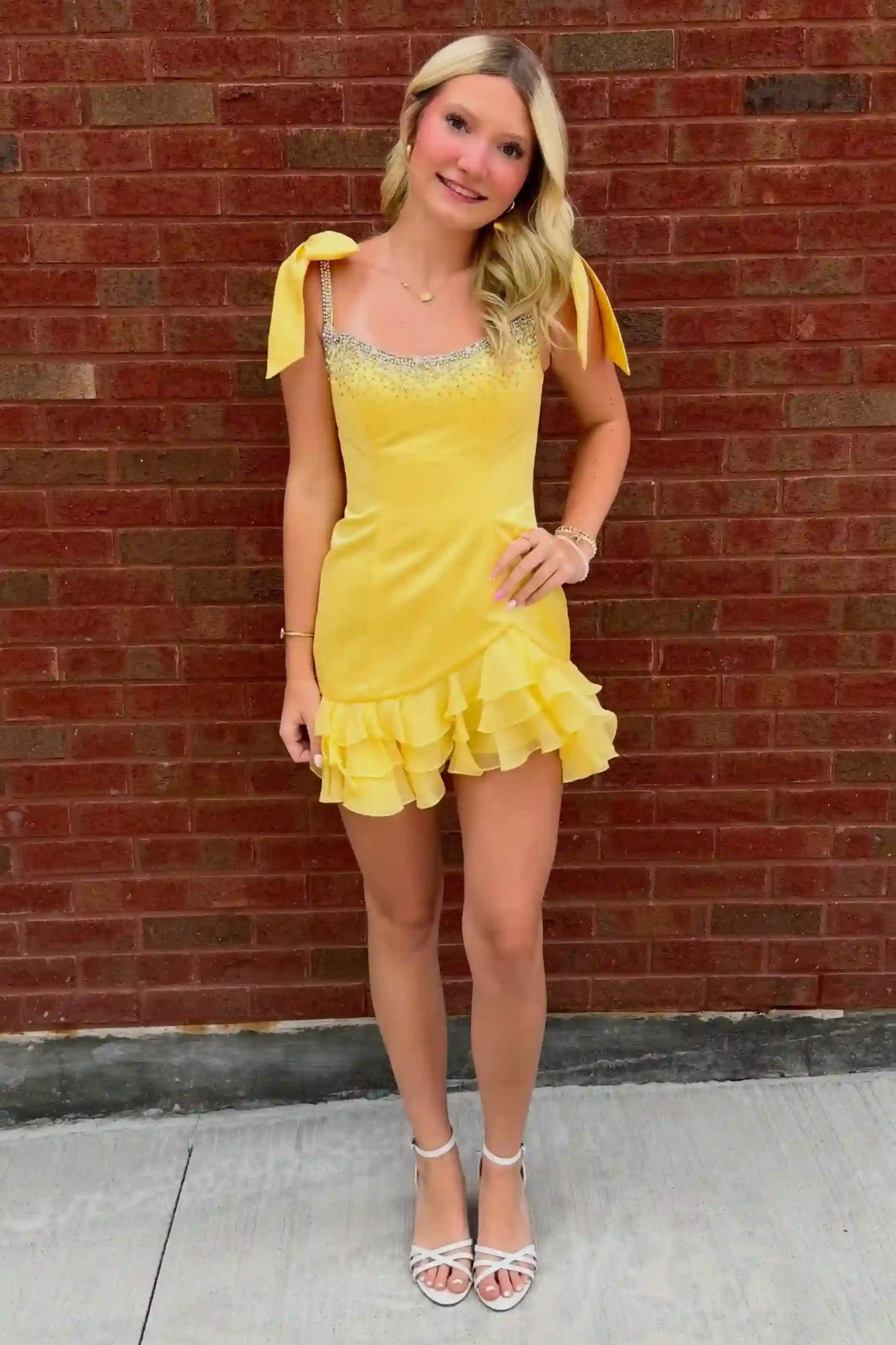 A blonde woman wearing a bright yellow mini dress with ruffled trim and bow details on the shoulders, standing against a red brick wall.